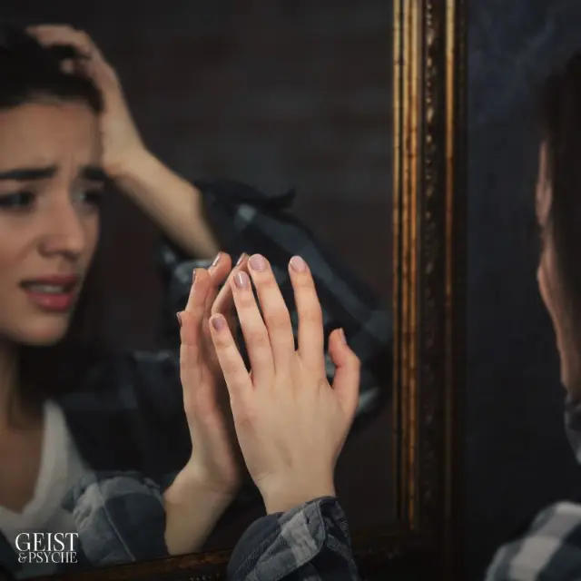 Eine Frau schaut besorgt in einen Spiegel und berührt ihr Spiegelbild mit der Hand, was Symptome fehlender Selbstliebe verdeutlicht. Der Ausdruck von Angst und Unsicherheit spiegelt die emotionalen Folgen dieser Problematik wider.